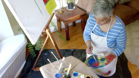 Senior-biracial-woman-wearing-apron-and-mixing-colours-on-palette-at-home,-slow-motion