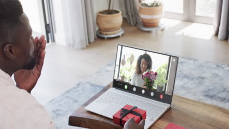 African-american-man-holding-red-gift-using-laptop-with-african-american-woman-and-flowers-on-screen