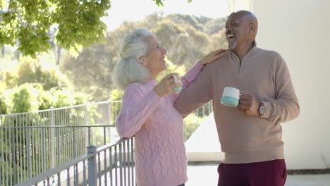Glückliches-älteres,-Gemischtrassiges-Paar,-Das-Sich-Zu-Hause-Auf-Dem-Balkon-Umarmt-Und-Kaffee-Trinkt,-Zeitlupe