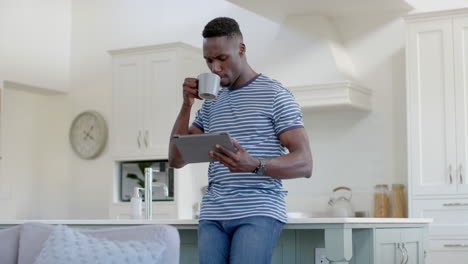 Happy-african-american-man-drinking-coffee-and-using-tablet-in-sunny-kitchen,-slow-motion