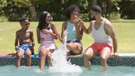 Happy-african-american-parents,-son-and-daughter-sitting-in-sun-splashing,-feet-in-pool,-slow-motion