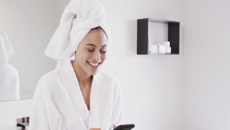 Portrait-of-happy-biracial-woman-with-towel-on-head-using-smartphone-in-bathroom,-slow-motion