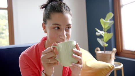 Portrait-of-happy-biracial-teenage-girl-sitting-at-home-drinking-tea,-copy-space,-slow-motion