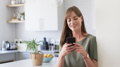 Middle-aged-Caucasian-woman-texts-on-her-phone-in-a-modern-kitchen