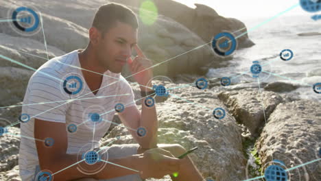 Animation-of-network-of-connections-with-icons-over-biracial-man-using-smartphone-by-sea