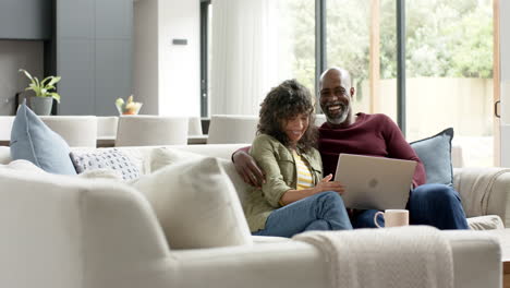 Glückliches-Biracial-Paar,-Das-Auf-Der-Couch-Sitzt-Und-Zu-Hause-Einen-Laptop-Benutzt,-Zeitlupe