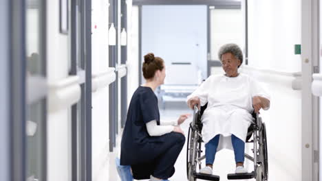 Diverse-female-doctor-talking-with-senior-woman-in-wheelchair-in-hospital-corridor,-slow-motion