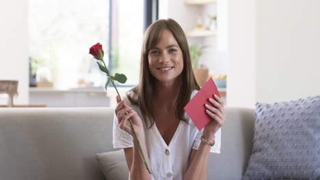 Middle-aged-Caucasian-woman-smiles-holding-a-rose-and-card-at-home-on-video-call