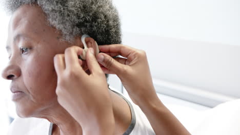 African-american-female-doctor-applying-hearing-aid-to-senior-woman-in-hospital-room,-slow-motion
