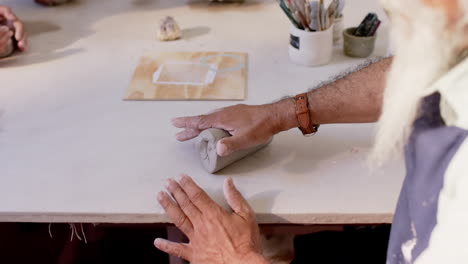 Focused-biracial-potter-with-long-beard-shaping-clay-with-hands-in-pottery-studio,-slow-motion