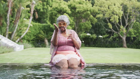 Happy-senior-african-american-woman-waving-by-pool-with-cocktail,-copy-space,-slow-motion