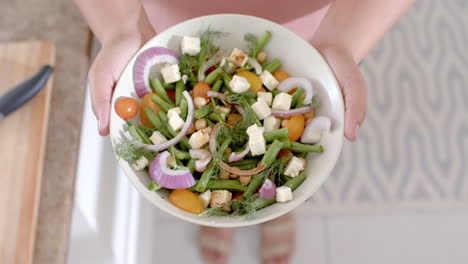 Overhead-hands-of-biracial-woman-holding-bowl-of-feta-salad-in-kitchen,-copy-space,-slow-motion