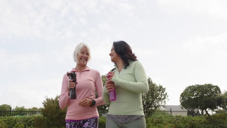 Zwei-Unterschiedliche-ältere-Frauen,-Die-Mit-Wasserflaschen-Spazieren-Gehen-Und-An-Einem-Sonnigen-Tag-Diskutieren,-Zeitlupe,-Kopierraum