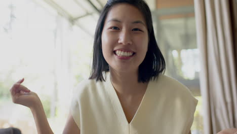 Mujer-Asiática-Feliz-Teniendo-Videollamada-Y-Sonriendo-En-Una-Sala-De-Estar-Soleada,-Cámara-Lenta