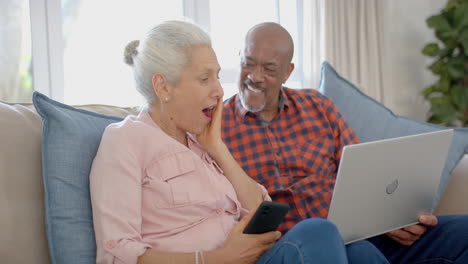 Happy-senior-biracial-couple-sitting-on-couch-and-using-laptop-at-home,-slow-motion