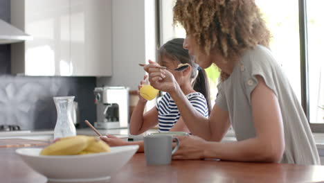 Biracial-mother-and-daughter-eating-breakfast-cereal-in-sunny-kitchen,-copy-space,-slow-motion