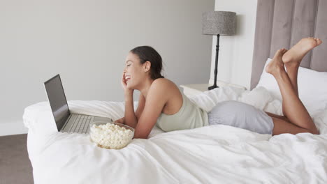Happy-biracial-woman-lying-on-bed-using-laptop-and-eating-popcorn,-slow-motion
