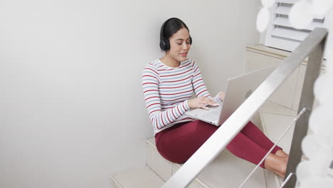 Feliz-Mujer-Birracial-Sentada-En-Las-Escaleras-Usando-Auriculares-Y-Computadora-Portátil,-Cámara-Lenta