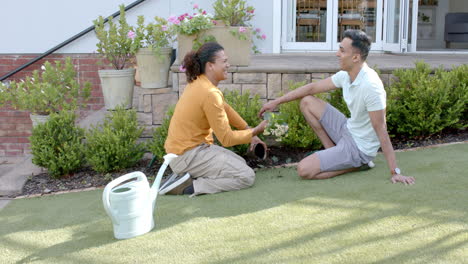 Happy-diverse-gay-male-couple-gardening-and-talking-kneeling-in-sunny-garden,-slow-motion