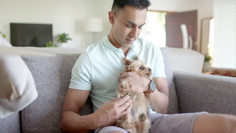 Happy-biracial-man-sitting-on-sofa,-petting-pet-yorkshire-terrier-dog-and-smiling,-slow-motion