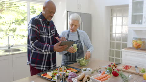 Happy-diverse-senior-couple-preparing-vegetables,-using-tablet-in-kitchen,-copy-space,-slow-motion