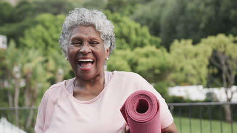 Portrait-of-happy-senior-african-american-woman-holding-yoga-mat,-laughing-on-balcony,-slow-motion
