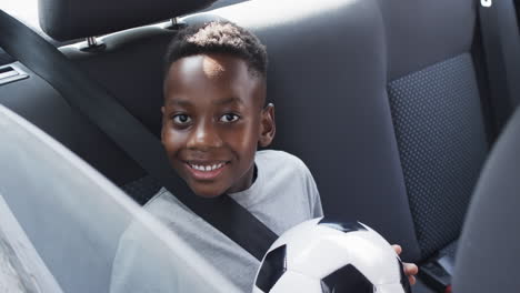 African-American-boy-smiles-in-the-backseat-of-a-car,-holding-a-soccer-ball
