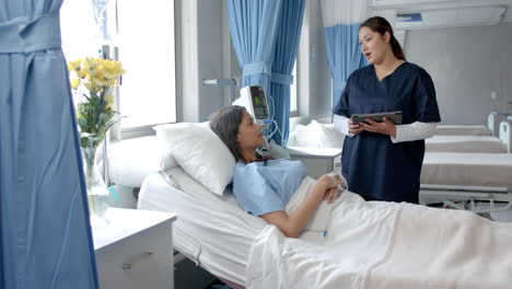 Diverse-female-patient-in-hospital-bed-and-doctor-with-tablet-talking,-copy-space,-slow-motion