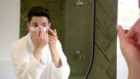 Focused-biracial-man-applying-under-eye-masks-looking-in-bathroom-mirror,-slow-motion