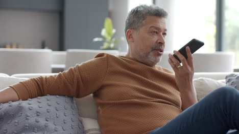 Älterer-Mann-Mit-Gemischter-Abstammung,-Der-Auf-Dem-Smartphone-Spricht-Und-Auf-Der-Couch-Im-Wohnzimmer-Sitzt,-Zeitlupe