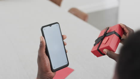 African-American-man-holding-a-smartphone-and-a-gift-box-at-home-with-copy-space,-on-a-video-call