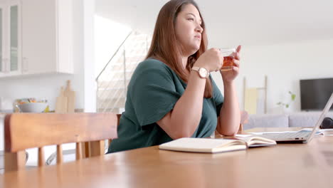 Thoughtful-plus-size-biracial-woman-drinking-tea-and-using-laptop,-working-from-home,-slow-motion