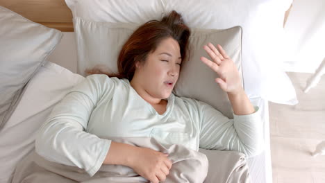 Happy-plus-size-biracial-woman-waking-up-in-bed,-stretching-and-yawning-in-the-morning,-slow-motion