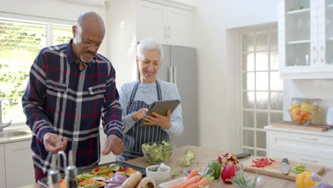 Feliz-Pareja-De-Ancianos-Diversos-Preparando-Verduras,-Usando-Tableta-En-La-Cocina,-Espacio-Para-Copiar,-Cámara-Lenta