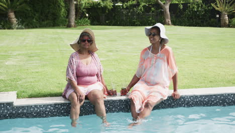 Glückliche-ältere-Afroamerikanische-Freundinnen-In-Sonnenhüten,-Die-Mit-Getränken-Am-Pool-Sitzen,-Zeitlupe