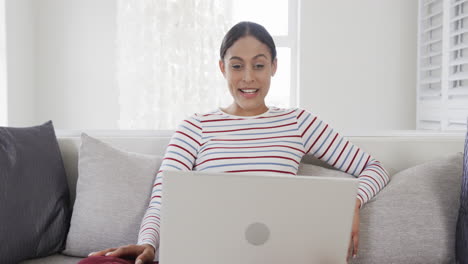 Feliz-Mujer-Birracial-Sentada-En-Un-Sofá-Usando-Una-Computadora-Portátil-Para-Videollamadas,-Cámara-Lenta