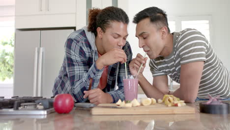 Happy-diverse-gay-male-couple-sharing-fruit-smoothie-drinking-with-straws-in-kitchen,-slow-motion