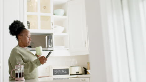 Glückliche-Afroamerikanische-Seniorin,-Die-Kaffee-Trinkt,-Ihr-Smartphone-In-Der-Sonnigen-Küche-Benutzt,-Zeitlupe