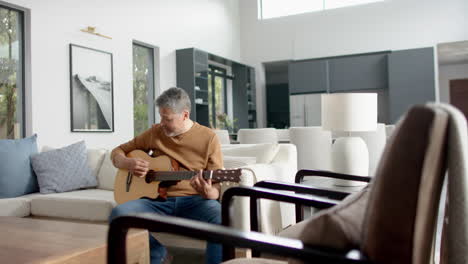 Älterer-Mann-Mit-Gemischter-Abstammung,-Der-Zu-Hause-Auf-Der-Couch-Sitzt-Und-Gitarre-Spielt,-Kopierraum,-Zeitlupe