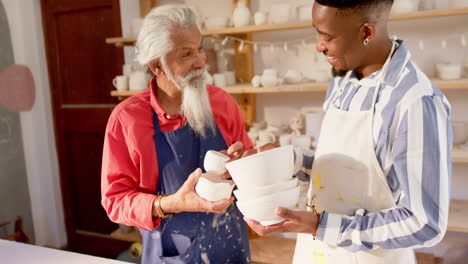 Happy-diverse-male-potters-holding-bowls-and-discussing-in-pottery-studio,-slow-motion