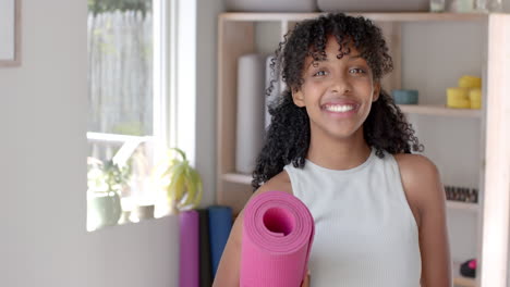 Portrait-of-happy-biracial-fitness-teenage-girl-holding-yoga-mat-in-white-room,-slow-motion
