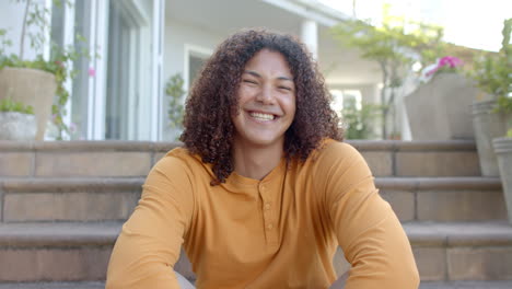Portrait-of-happy-biracial-man-with-long-hair-sitting-on-steps-smiling-in-sunny-garden,-slow-motion