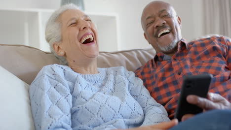Happy-senior-biracial-couple-sitting-on-couch-and-using-smartphone-at-home,-slow-motion