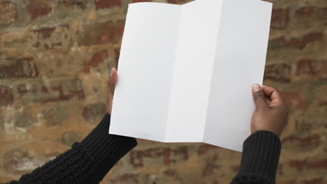 Video-of-hand-of-african-american-man-holding-white-blank-page,-copy-space-over-brick-wall