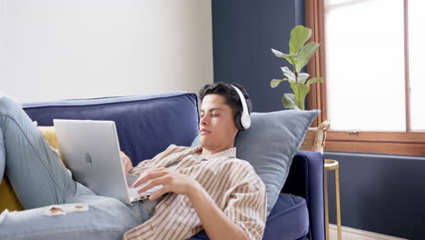 Focused-biracial-man-wearing-headphones-lying-on-couch-using-laptop-at-home,-copy-space,-slow-motion
