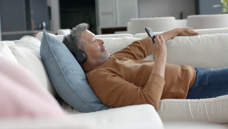 Senior-biracial-man-on-couch-in-headphones-listening-to-music-on-smartphone,-copy-space,-slow-motion