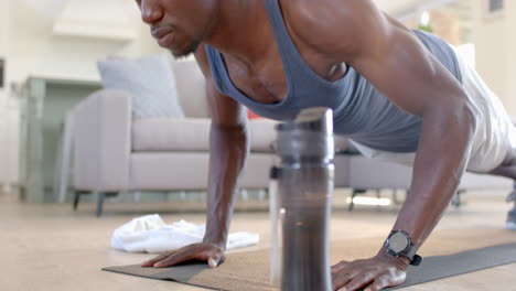 Focused-african-american-man-doing-push-ups-next-to-water-bottle-in-sunny-living-room,-slow-motion