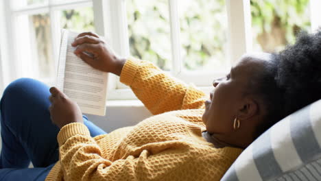 Happy-african-american-senior-woman-lying,-reading-book-and-smiling-in-sunny-room,-slow-motion