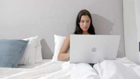 Focused-biracial-teenage-girl-sitting-in-bed-in-the-morning-using-laptop,-copy-space,-slow-motion
