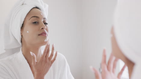 Biracial-woman-applying-face-cream-looking-in-mirror-in-bathroom,-slow-motion
